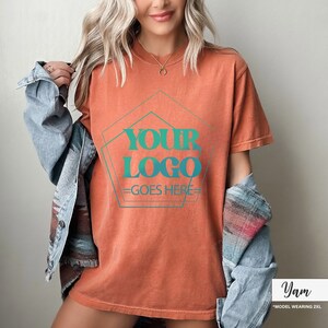 May include: Burnt orange t-shirt with the text "YOUR LOGO GOES HERE" in teal. The text is inside a teal pentagon shape. The model is wearing a denim jacket over the t-shirt. The model is wearing a size 2XL.
