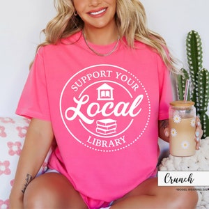 May include: A bright pink t-shirt with the text "SUPPORT YOUR LOCAL LIBRARY" in a white circular design. The design includes a building and books. A glass with daisy decals and a straw is visible.