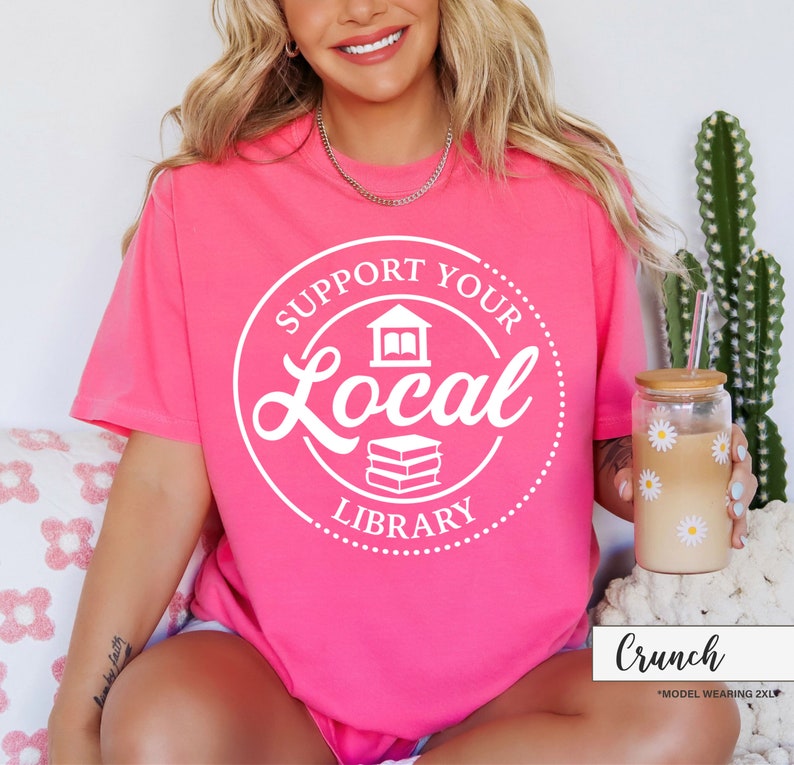 May include: A bright pink t-shirt with the text "SUPPORT YOUR LOCAL LIBRARY" in a white circular design. The design includes a building and books. A glass with daisy decals and a straw is visible.