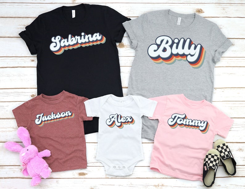 May include: Five t-shirts with retro-style names printed on them. The shirts are in different colours and sizes. The names are "Sabrina", "Billy", "Jackson", "Alex", and "Tommy".