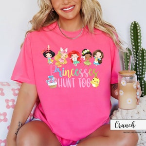 May include: A bright pink t-shirt with the text "Princesses Hunt Too" and cartoon illustrations of princesses and Easter-themed elements. The shirt features a crew neck and short sleeves.