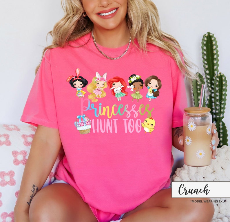 May include: A bright pink t-shirt with the text "Princesses Hunt Too" and cartoon illustrations of princesses and Easter-themed elements. The shirt features a crew neck and short sleeves.