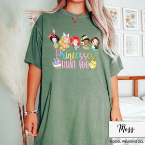 May include: Moss green t-shirt featuring a graphic of cartoon princesses and the text "Princesses Hunt Too." The design includes a basket of eggs and a chick. The shirt is a comfortable, oversized fit.