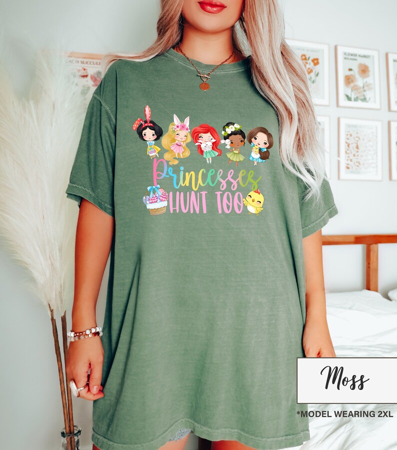 May include: Moss green t-shirt featuring a graphic of cartoon princesses and the text "Princesses Hunt Too." The design includes a basket of eggs and a chick. The shirt is a comfortable, oversized fit.