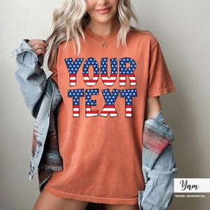 May include: Orange-red t-shirt with the text "YOUR TEXT" in a patriotic design featuring blue stars and red and white stripes. The model is wearing a denim jacket. The shirt is a 2XL.