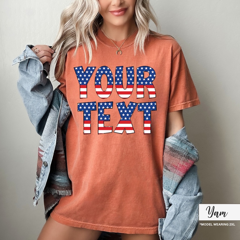 May include: Orange-red t-shirt with the text "YOUR TEXT" in a patriotic design featuring blue stars and red and white stripes. The model is wearing a denim jacket. The shirt is a 2XL.