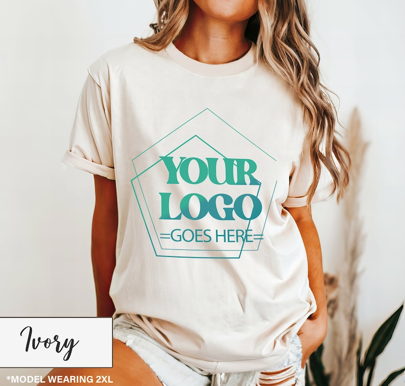 May include: Ivory-colored t-shirt with a crew neck and rolled-up sleeves. The shirt features a teal and green geometric design with the text "YOUR LOGO GOES HERE." The bottom left corner displays the word "Ivory" in a white box.