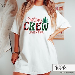 Może przedstawiać: Biała koszulka z napisem "Christmas Crew" w kolorze czerwonym i zielonym. Słowo "Crew" ma wz&oacute;r w kratę. Koszulka posiada r&oacute;wnież zieloną grafikę choinki oraz napis "Custom Name".