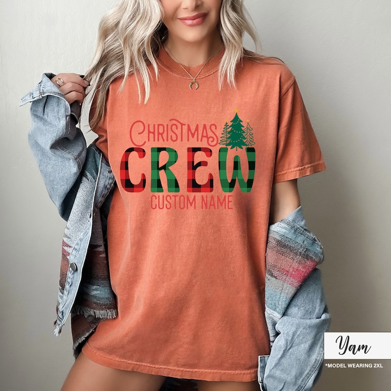 Może przedstawiać: Pomarańczowa koszulka z napisem "Christmas Crew" w czerwono-zieloną kratę. Koszulka posiada grafikę choinki i napis "Custom Name" pod słowem "Crew".
