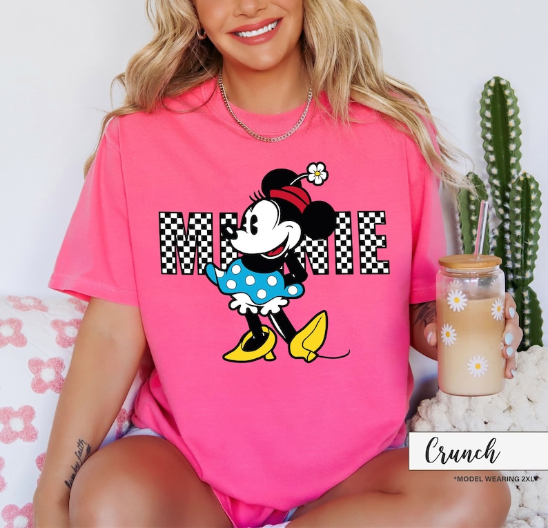 May include: A bright pink t-shirt featuring a graphic of Minnie Mouse. The word "MINNIE" is displayed in a checkered pattern. Minnie Mouse is wearing a blue polka dot dress, yellow shoes, and a red hat with a flower. The shirt is labeled "Crunch".