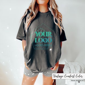May include: Dark gray t-shirt with the text "YOUR LOGO GOES HERE" in a teal and turquoise design. The shirt is a vintage comfort color and is a 2XL size.