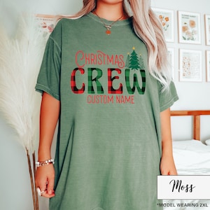 Może przedstawiać: Zielona koszulka w kolorze mchu z napisem "Christmas Crew" w czerwonych i kratkowanych literach. Zielona grafika choinki znajduje się po prawej stronie. Słowa "Custom Name" znajdują się poniżej. Modelka nosi rozmiar 2XL.