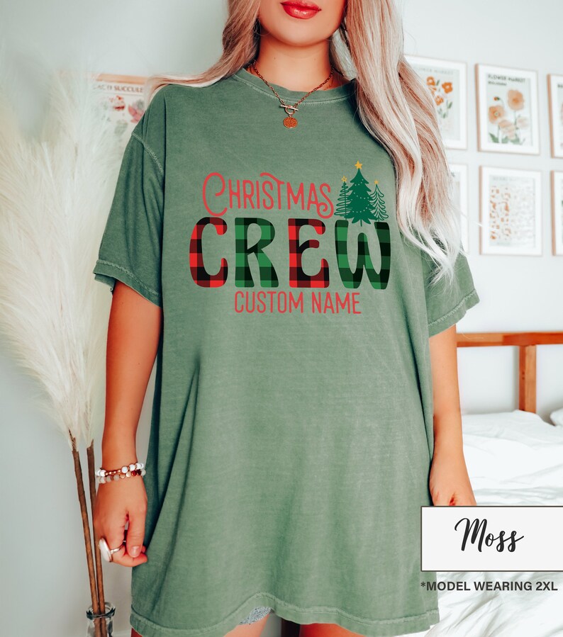 Może przedstawiać: Zielona koszulka w kolorze mchu z napisem "Christmas Crew" w czerwonych i kratkowanych literach. Zielona grafika choinki znajduje się po prawej stronie. Słowa "Custom Name" znajdują się poniżej. Modelka nosi rozmiar 2XL.