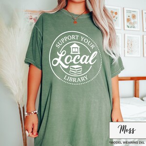 May include: Moss green t-shirt with a white circular graphic that reads "Support Your Local Library". The graphic includes a house and a stack of books. The shirt is a relaxed fit and has short sleeves. The model is wearing a 2XL size.