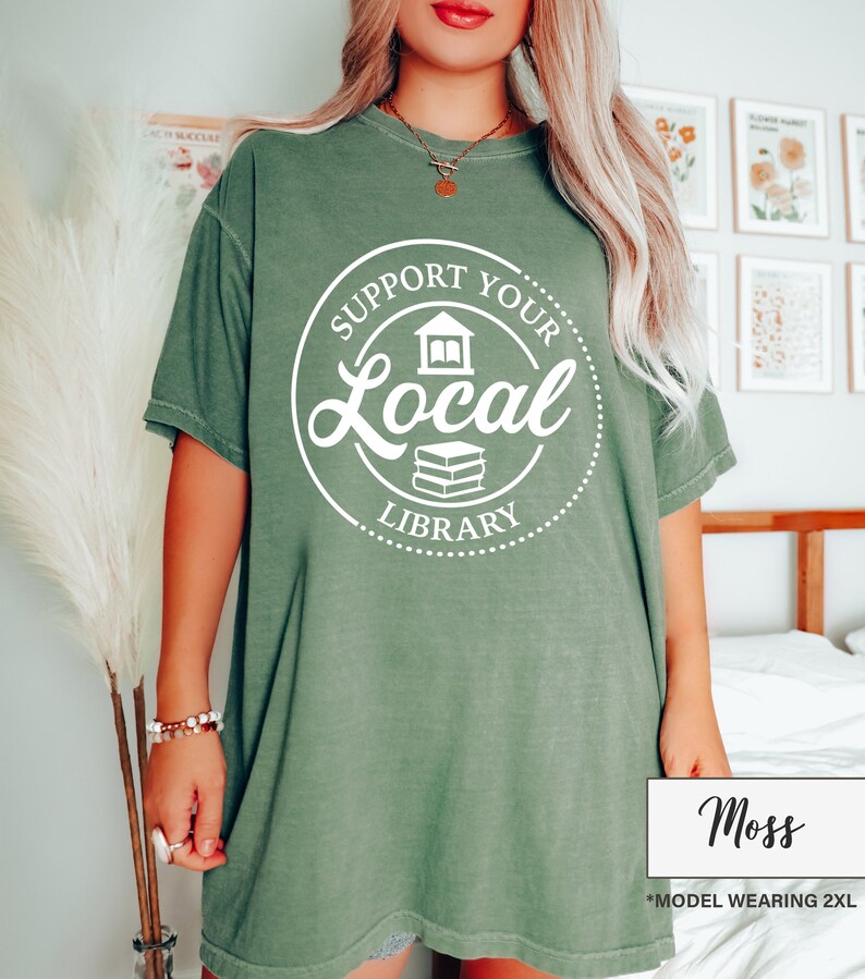 May include: Moss green t-shirt with a white circular graphic that reads "Support Your Local Library". The graphic includes a house and a stack of books. The shirt is a relaxed fit and has short sleeves. The model is wearing a 2XL size.