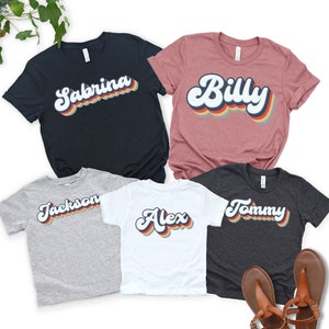 May include: Five t-shirts with retro-style font names printed on them. The names are Sabrina, Billy, Jackson, Alex, and Tommy. The shirts are in various colours, including black, pink, grey, white, and dark grey.
