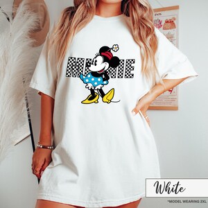 May include: White t-shirt featuring a Minnie Mouse graphic. The graphic includes the word "MINNIE" in a black and white checkered pattern, along with a cartoon illustration of Minnie Mouse in a blue polka dot dress and yellow shoes.