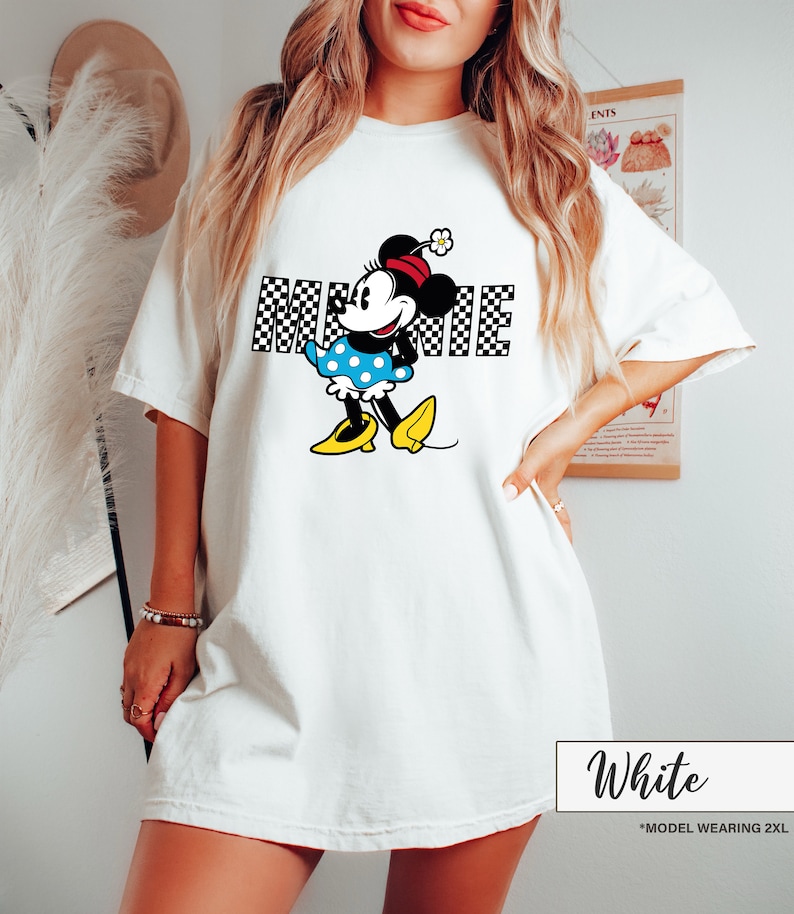 May include: White t-shirt featuring a Minnie Mouse graphic. The graphic includes the word "MINNIE" in a black and white checkered pattern, along with a cartoon illustration of Minnie Mouse in a blue polka dot dress and yellow shoes.