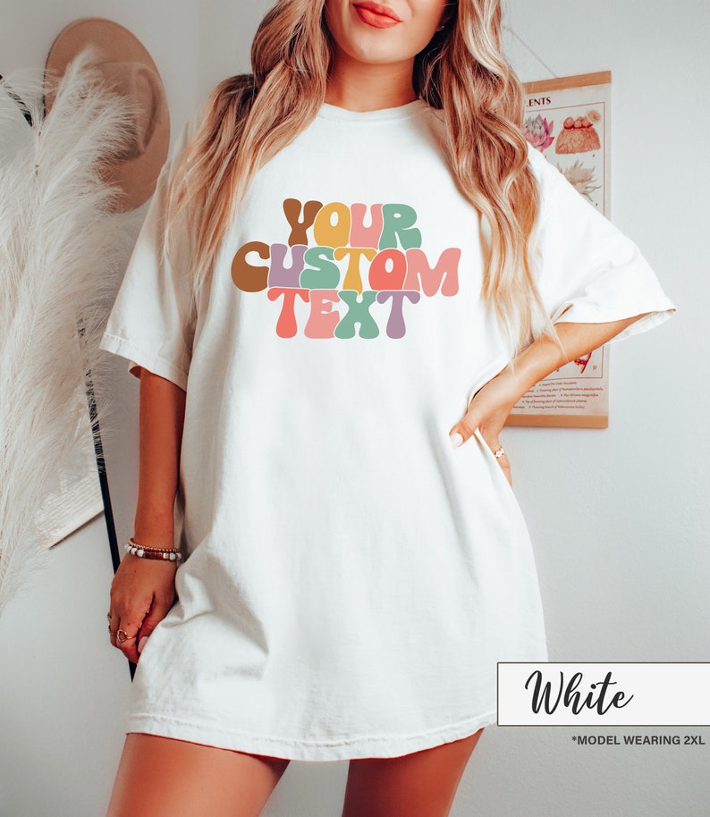 Op de afbeelding: Wit oversized T-shirt met de tekst "YOUR CUSTOM TEXT" in kleurrijke, retro-stijl letters.
