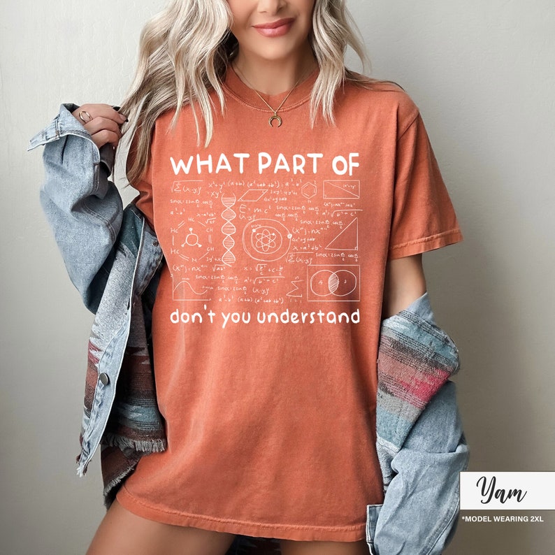May include: Burnt orange t-shirt with the text "WHAT PART OF don't you understand" in white, along with scientific diagrams. The shirt is a relaxed fit and has short sleeves. The model is wearing a denim jacket.