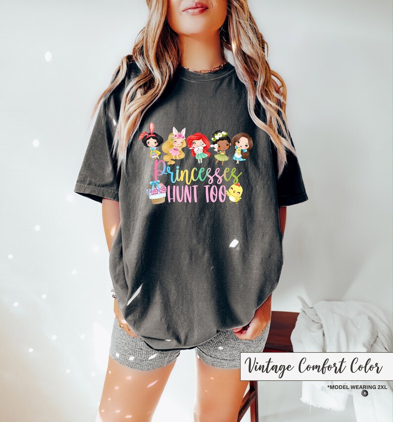 May include: Dark gray t-shirt featuring a colorful graphic of cartoon princesses and the text "Princesses Hunt Too." The shirt is a vintage comfort color and has a relaxed fit. The model is wearing a 2XL size.