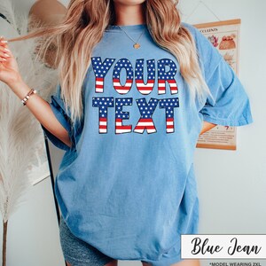 May include: A light blue t-shirt with the text "YOUR TEXT" in a patriotic design of red and white stripes and blue stars. The shirt is a relaxed fit and has short sleeves. The words "Blue Jean" are written in cursive in the bottom right corner.