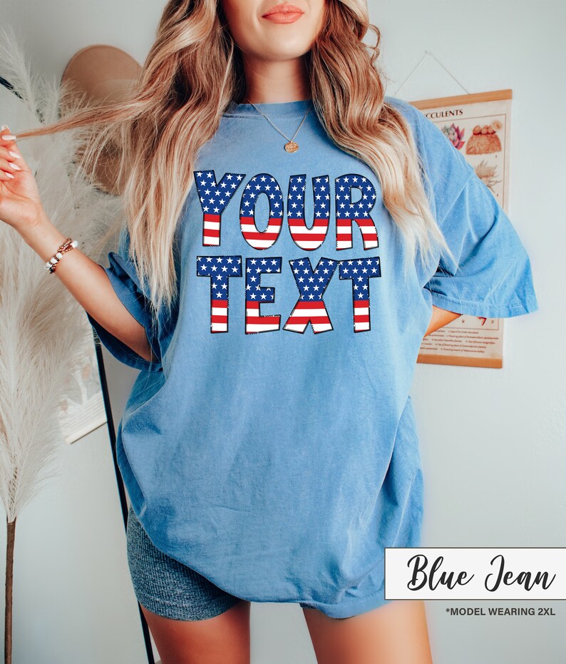 May include: A light blue t-shirt with the text "YOUR TEXT" in a patriotic design of red and white stripes and blue stars. The shirt is a relaxed fit and has short sleeves. The words "Blue Jean" are written in cursive in the bottom right corner.