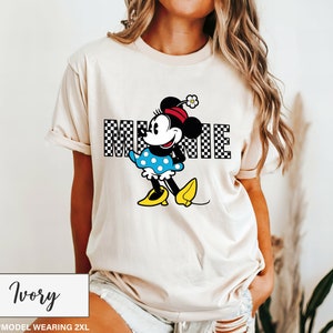 May include: Ivory-colored t-shirt featuring a Minnie Mouse graphic. The design includes the word "MINNIE" in a checkered pattern, with Minnie Mouse wearing a red bow, blue polka-dot dress, and yellow shoes. The shirt is a 2XL size.