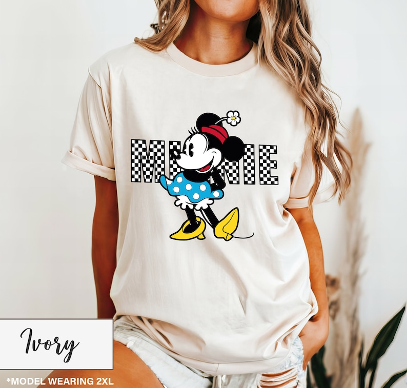 May include: Ivory-colored t-shirt featuring a Minnie Mouse graphic. The design includes the word "MINNIE" in a checkered pattern, with Minnie Mouse wearing a red bow, blue polka-dot dress, and yellow shoes. The shirt is a 2XL size.