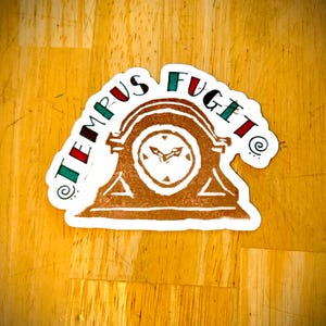 May include: A sticker featuring the Latin phrase "TEMPUS FUGIT" in red and green, encircling a brown clock illustration. The clock has a white face and a triangular base. The sticker is placed on a wooden surface.