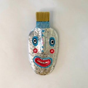 Pode incluir: Uma máscara prateada com um rosto pintado. A máscara tem listras azuis e brancas na parte superior, lábios vermelhos, bochechas cor-de-rosa e olhos pretos.