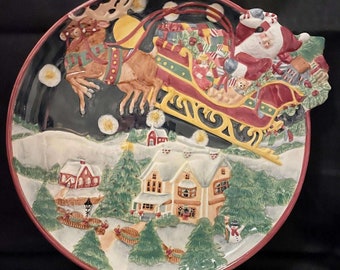 Może przedstawiać: Ceramiczny talerz świąteczny z trójwymiarową postacią Świętego Mikołaja w saniach ciągniętych przez renifery, lecących nad śnieżną wioską. Talerz ma ciemnoniebieskie tło z białymi płatkami śniegu i gwiazdami.