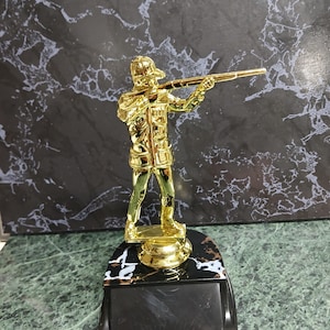 May include: A gold-colored trophy featuring a person holding a rifle. The trophy has a black base with a white marble design.