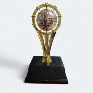 May include: A gold-colored trophy with a mirrored disco ball at the top, surrounded by a ring of stars. The trophy sits on a black base with a gold stem. The trophy is a symbol of achievement.