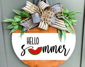 May include: A round wooden sign with a white background and the text "Hello Summer" in black script. A red watermelon slice with black seeds is between the words "Hello" and "Summer". The sign is decorated with a burlap and blue striped bow and green leaves.