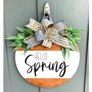 May include: A round wooden door hanger with a white background and the text "HELLO Spring" in black script. The hanger is decorated with a burlap bow and green leaves.