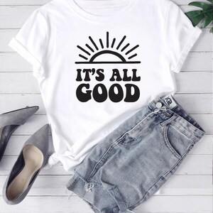 Può includere: Maglietta bianca con una grafica nera di un sole e il testo "IT'S ALL GOOD" in un font retrò.
