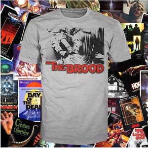 Peut inclure: Un t-shirt gris avec une image en noir et blanc d'une femme et d'un enfant. Le texte "THE BROOD" est imprimé en lettres rouges sous l'image.