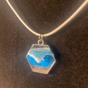 May include: A hexagonal pendant necklace with a silver-coloured frame. The pendant features a blue and white design, resembling ocean waves, set in clear resin. The necklace has a white cord.