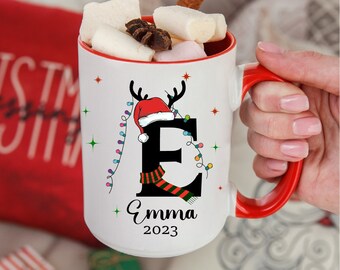 Könnte beinhalten: Weißer Keramikbecher mit rotem Innenraum und Henkel, verziert mit einem weihnachtlichen Buchstaben "E" mit Weihnachtsmütze und Rentiergeweih. Der Becher ist mit Marshmallows und einem Zimtstange gefüllt. Der Name "Emma" und das Jahr "2023" sind aufgedruckt.