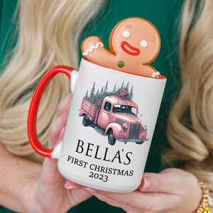 Könnte beinhalten: Weißer Keramikbecher mit roter Innenseite und Henkel, mit einer Illustration eines rosa Oldtimer-Lastwagens, der einen Weihnachtsbaum transportiert. Der Becher trägt den Text "BELLA'S FIRST CHRISTMAS 2023". Ein Lebkuchenmann-Keks sitzt am Rand.