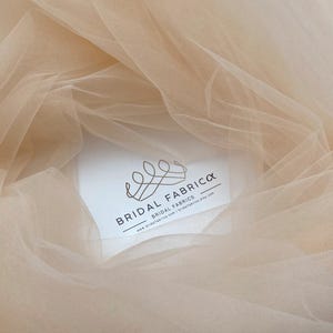 May include: A close-up of a pale peach tulle fabric. The fabric is soft and sheer, with a delicate texture. The fabric is draped over a white card with the text "Bridal Fabrica Bridal Fabrics www.bridalfabrica.com | bridalfabrica.etsy.com"