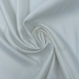 May include: White satin fabric, smooth and silky, with a soft drape. The fabric is folded and arranged in a way that shows its texture and flow.