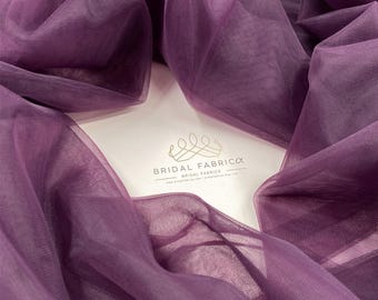 May include: Purple tulle fabric, soft and sheer, with a delicate texture. The fabric is draped and folded, showcasing its flowy nature. The text "BRIDAL FABRICCA BRIDAL FABRICS" is visible in the lower center of the image.