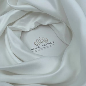 May include: White satin fabric, a smooth and luxurious material, draped in soft folds. The fabric is a popular choice for wedding gowns and other formal wear.