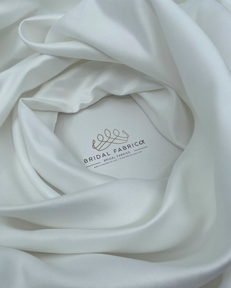 May include: White satin fabric, a smooth and luxurious material, draped in soft folds. The fabric is a popular choice for wedding gowns and other formal wear.