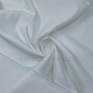 May include: White satin fabric with a smooth, silky texture. The fabric is folded and draped, showcasing its luxurious feel.