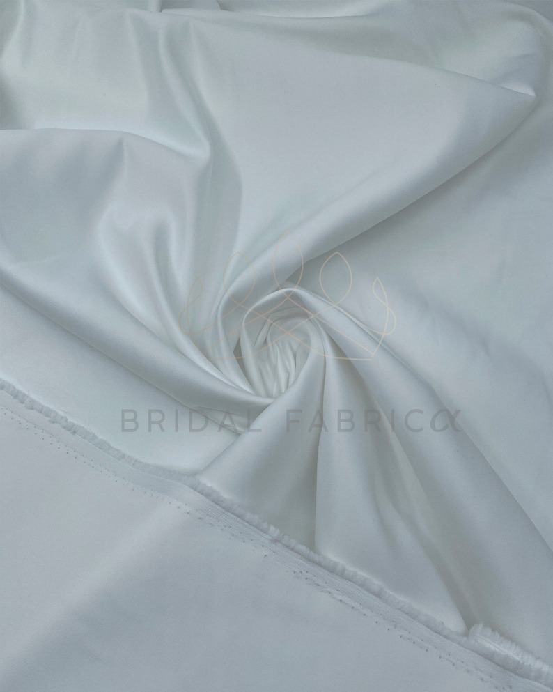 May include: White satin fabric with a smooth, silky texture. The fabric is folded and draped, showcasing its luxurious feel.