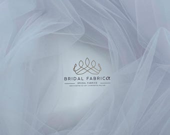 May include: White tulle fabric with a soft, delicate texture. The fabric is draped and folded, creating a flowing and ethereal look. The image includes the logo "Bridal Fabrica" with the tagline "Bridal Fabrics" and the website address "www.bridalfabrica.com | bridalfabrica.etsy.com".