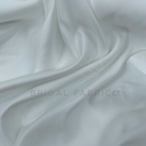 May include: White satin fabric, smooth and silky, with a subtle sheen. The fabric is draped and folded, showcasing its luxurious texture.